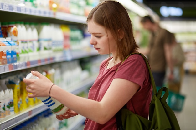 Foto di attraente giovane consumatore modello femminile con acconciatura bobbed, vestita in maglietta casual, si trova nel grande negozio, detiene una bottiglia di latte