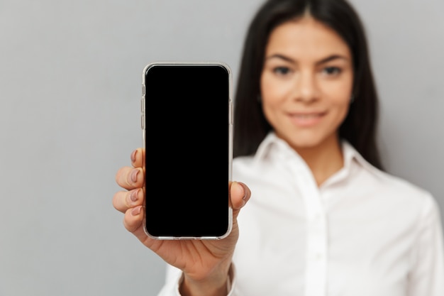 Foto di attraente giovane donna 30s in abbigliamento da ufficio in piedi con lo smartphone in mano e mostrando lo schermo nero del copyspace alla macchina fotografica, isolata sopra fondo grigio