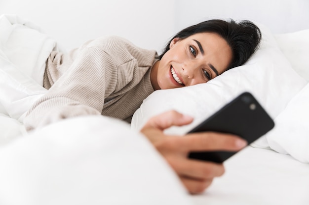 Foto di bella donna 30s utilizza lo smartphone, mentre giaceva a letto con lenzuola bianche a casa