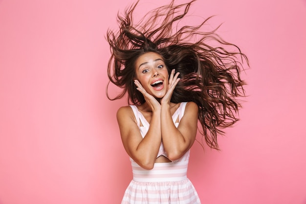 Foto di bella donna anni '20 che indossa un abito sorridente e scuotendo i suoi lunghi capelli castani, isolato sopra il muro rosa