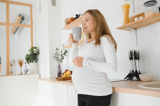 Foto di bella giovane donna bruna vestita casualmente in attesa di un bambino, che mangia il tè del mattino in una cucina moderna ed elegante, sorridente. Anticipazione, aspettativa, gravidanza e concetto di maternità