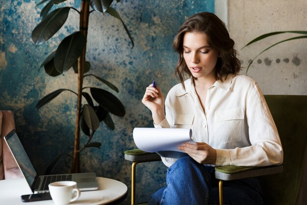 Foto di bella giovane donna seduta in un caffè al chiuso lavora con i documenti della holding del laptop.