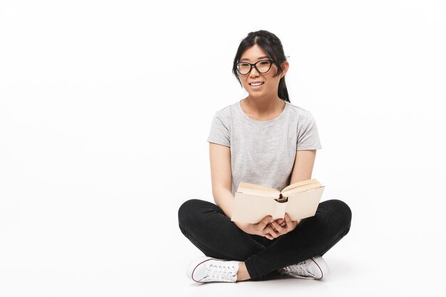 Foto di bella posa asiatica della giovane donna isolata sopra la lettura bianca del libro della tenuta della parete.