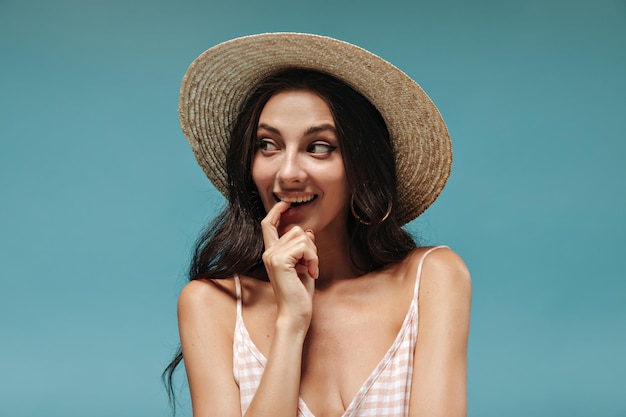 Foto di bella signora attraente con capelli neri ondulati in orecchini d'argento cappello alla moda e top rosa che distoglie lo sguardo e sorridente