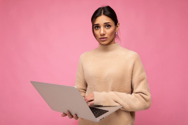 Foto di bella triste triste giovane donna bruna che indossa un maglione beige che tiene computer netbook digitando il testo sulla tastiera che guarda l'obbiettivo