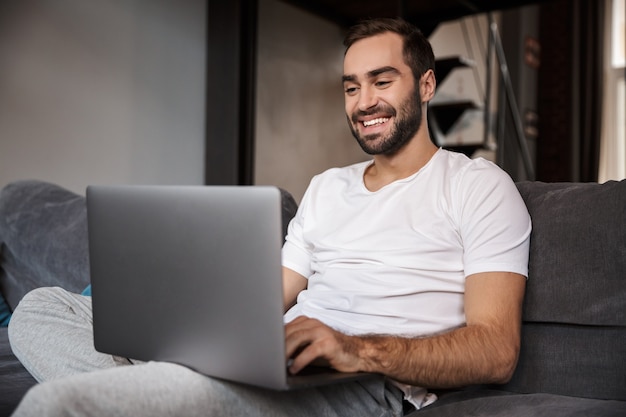 Foto di bruna scapolo 30s indossando t-shirt casual utilizzando laptop argento mentre era seduto sul divano nel soggiorno