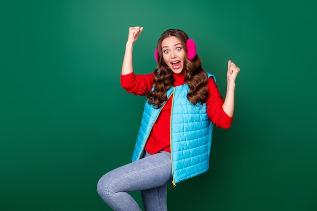 Foto di carina bella giovane donna bocca aperta pazzo sguardo stupito alzare i pugni ginocchio vincere squadra lotta con le palle di neve indossare jeans rosa scalda orecchie blu maglia felpa rossa isolato sfondo colore verde