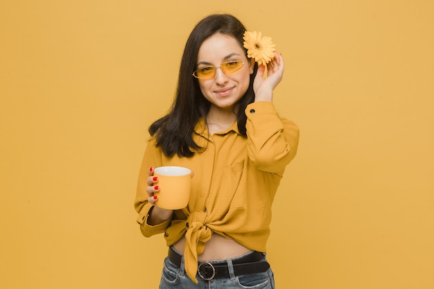 Foto di concetto di giovane donna in bicchieri e fiori nei capelli, detiene la bevanda, sentendosi tempo di primavera. Indossa maglietta gialla, sfondo di colore giallo isolato