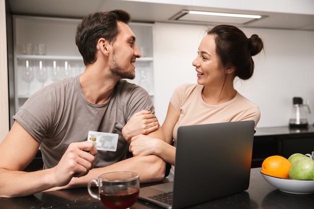 Foto di coppia allegra uomo e donna che utilizza computer portatile con carta di credito, mentre era seduto in cucina