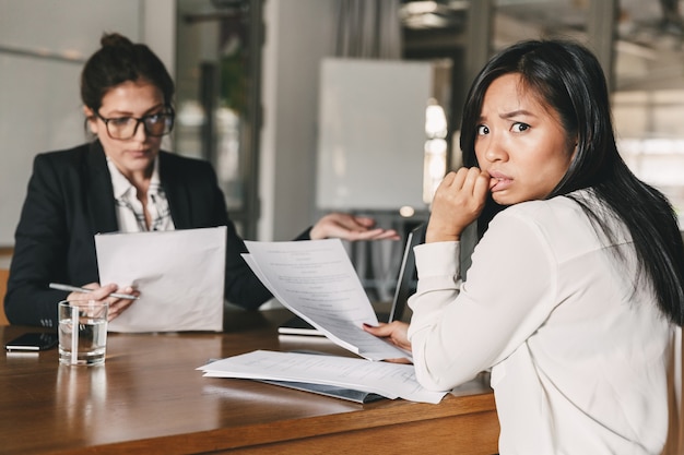 Foto di donna asiatica stressata che esprime il panico mentre è seduto al tavolo in ufficio e parla con un dipendente di sesso femminile, durante il colloquio di lavoro - concetto di affari, carriera e reclutamento