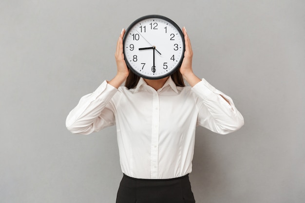 Foto di donna divertente in camicia bianca e gonna nera che copre il viso con un grande orologio rotondo, isolato sopra il muro grigio