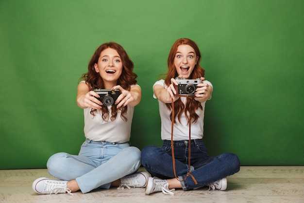 Foto di due ragazze 20s allegre rosse in jeans che si siedono sul pavimento con le gambe incrociate e dimostrando telecamere retrò, isolate su sfondo verde