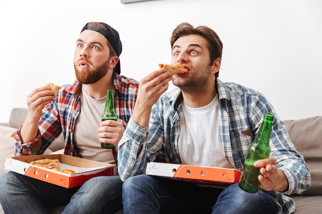 Foto di due ragazzi tesi in camicie casual che mangiano pizza e bevono birra, mentre guardano attentamente la partita di calcio a casa