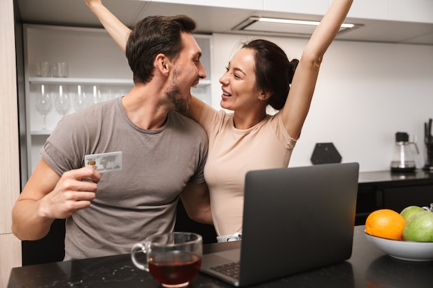 Foto di esultare coppia uomo e donna utilizzando laptop con carta di credito, mentre è seduto in cucina