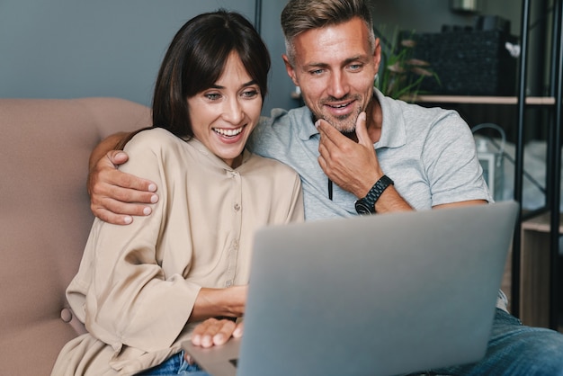 Foto di felice coppia caucasica uomo e donna che sorridono mentre guardano il laptop sul divano di casa