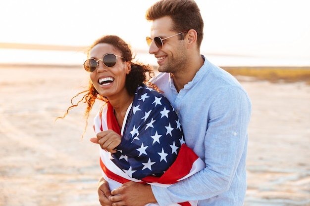 Foto di felice coppia multietnica uomo e donna avvolti nella bandiera americana che sorridono e si abbracciano insieme in riva al mare