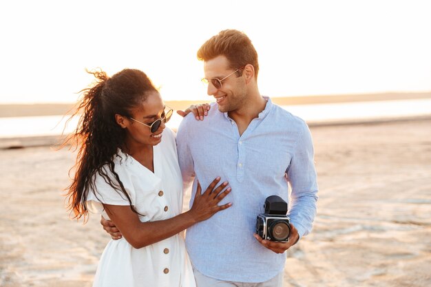 Foto di felice coppia multietnica uomo e donna che sorridono e si abbracciano insieme mentre si cammina in riva al mare con una fotocamera retrò