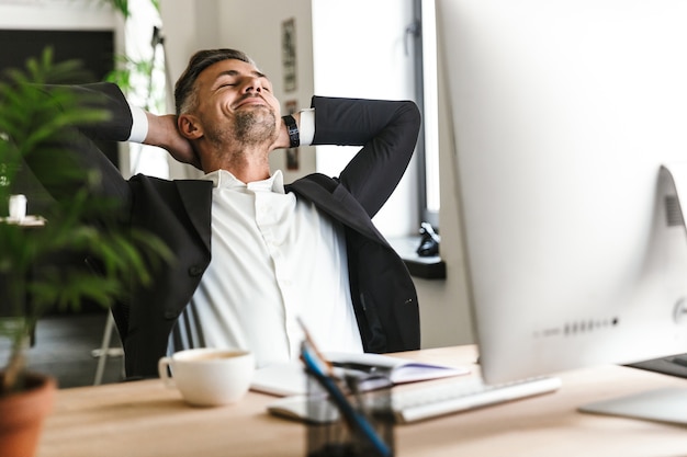 Foto di felice uomo d'affari 30s indossa tuta sorridente mentre era seduto alla scrivania e lavora al computer in ufficio