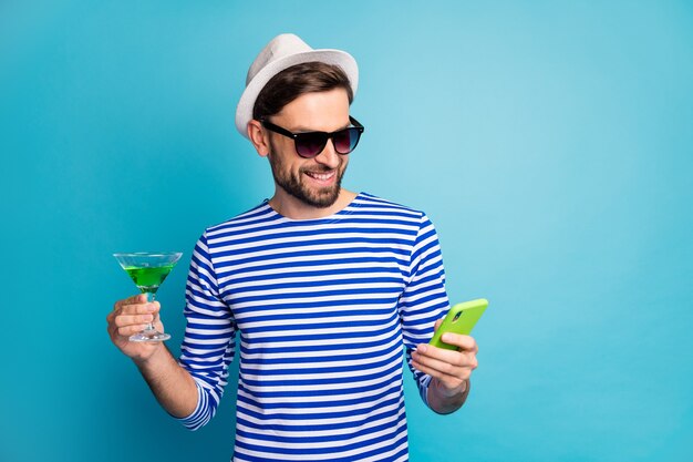 Foto di funky traveller guy navigazione telefono bere alcol verde cocktail all inclusive resort esotico indossare le specifiche del sole a strisce sailor shirt cap isolato di colore blu