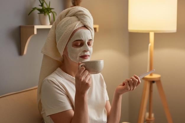 Foto di gioiosa giovane donna adulta soddisfatta con maschera cosmetologica sul viso seduta sulla tosse facendo manicure e annusando il caffè in tazza godendosi la mattina del fine settimana