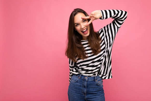 Foto di giovane bella donna castana felice accattivante sorridente positiva europea con emozioni sincere che indossa pullover a righe casual isolato su sfondo rosa con spazio di copia.