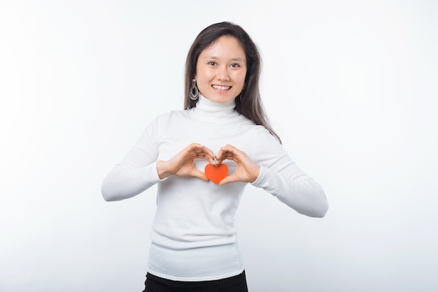 Foto di giovane donna asiatica affascinante che sorride e che tiene cuore di carta rosso sopra fondo bianco