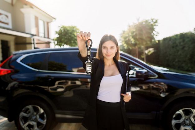 Foto di giovane donna caucasica felice che mostra chiave alla sua nuova automobile, stante davanti all'automobile nera all'aperto. Concetto per noleggio auto e acquisto. Concentrarsi sulla chiave