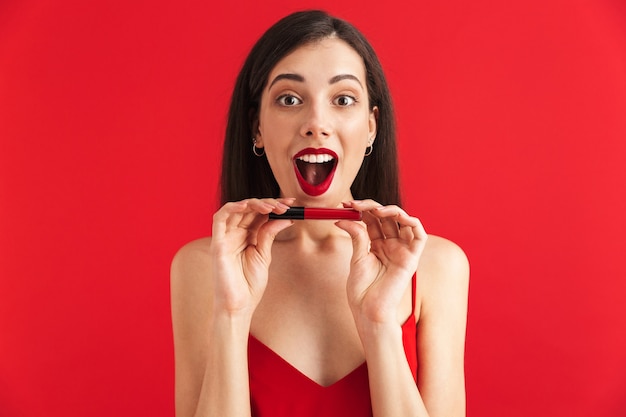 Foto di giovane donna emozionante felice che posa lucidalabbra isolato della tenuta che fa trucco.