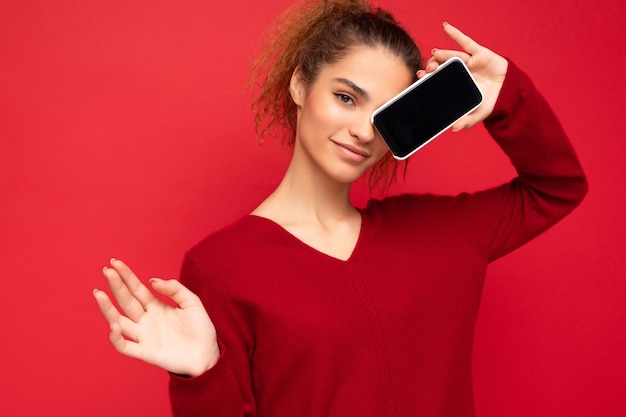 Foto di giovane femmina felice sexy che indossa un maglione rosso scuro isolato su sfondo rosso che tiene smartphone e che mostra lo schermo del telefono cellulare con lo spazio della copia per ritaglio che guarda l'obbiettivo.