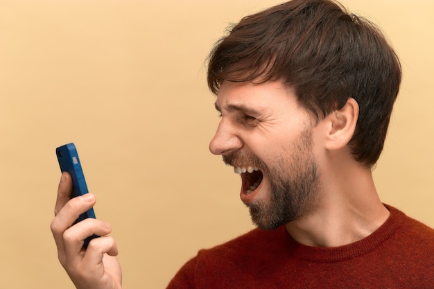 Foto di giovane uomo con la barba che indossa un maglione che urla di rabbia e shock, utilizzando il telefono cellulare, sentendosi frustrato per le informazioni sullo schermo, in posa contro il muro beige