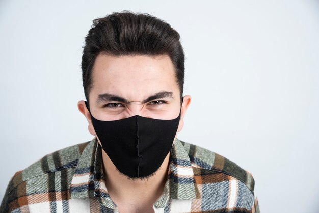 Foto di giovane uomo con maschera nera per protezione coronavirus in piedi sopra il muro bianco.