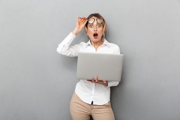 Foto di imprenditrice emotiva con gli occhiali in piedi e tenendo il computer portatile in ufficio, isolato