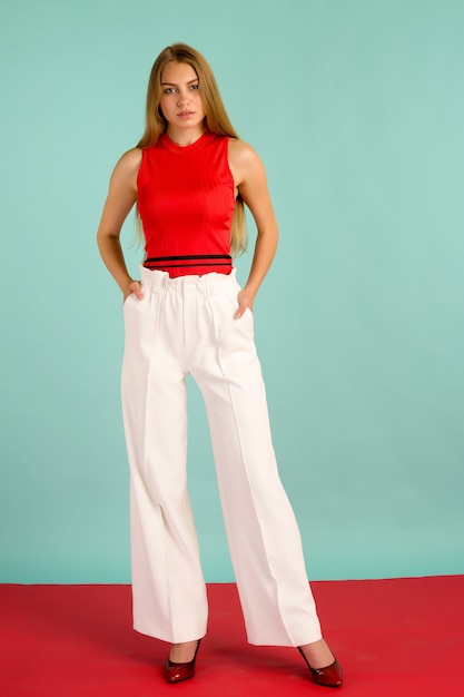 Foto di moda di una bella giovane donna elegante in una bella canotta rossa, pantaloni bianchi, tacchi alti in posa su sfondo blu.