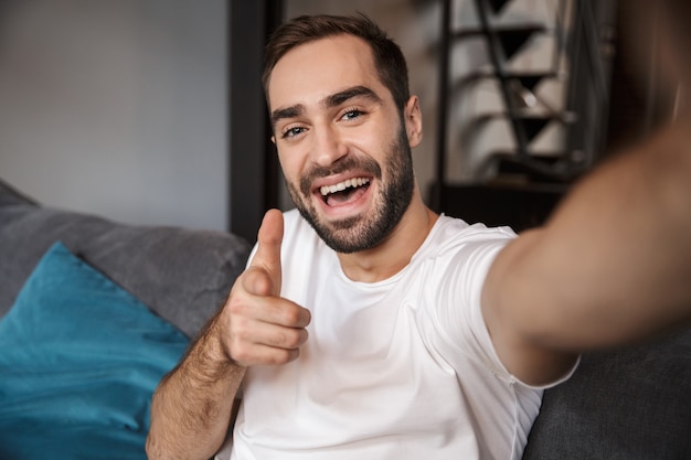 Foto di scapolo felice anni '30 che indossa una maglietta casual tenendo e prendendo selfie sul cellulare mentre è seduto sul divano nel soggiorno