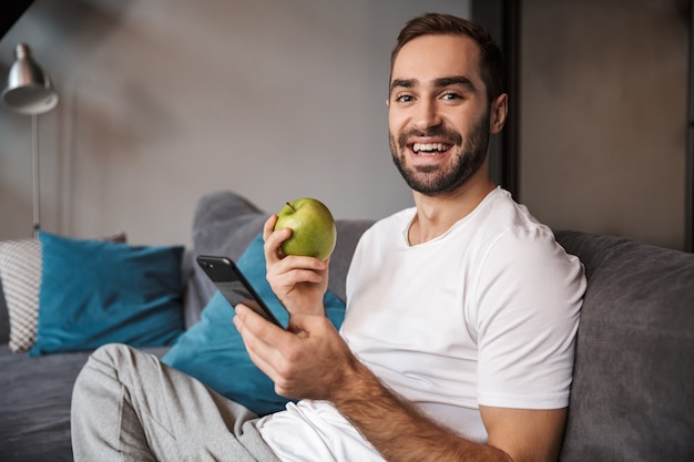 Foto di scapolo non rasato 30 anni che indossa una maglietta casual che tiene e utilizza il cellulare mentre era seduto sul divano nel soggiorno