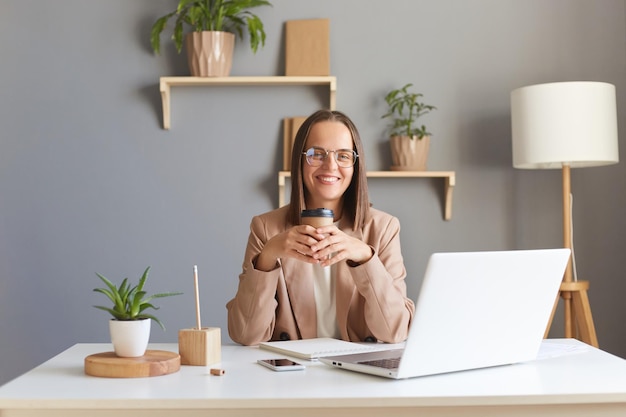 Foto di sorridente gioiosa allegra donna caucasica che indossa una giacca beige in posa in ufficio seduto sul posto di lavoro con pausa bevendo caffè godendosi il riposo durante il lavoro