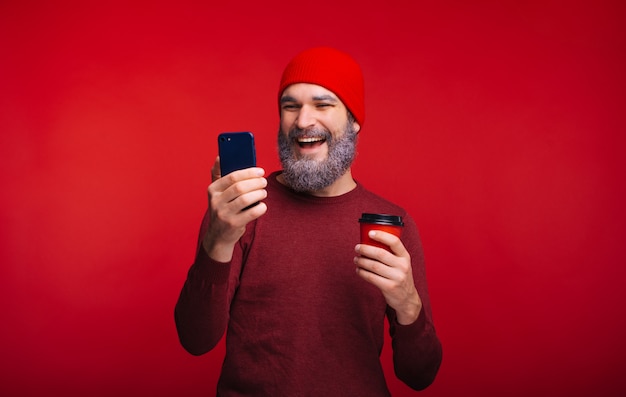 Foto di sorridente uomo barbuto in rosso guardando il telefono e tenendo la tazza di caffè