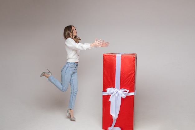 Foto di stock a figura intera di ragazza allegra che salta con i tacchi con le braccia tese con un regalo gigante in carta rossa con fiocco bianco sul muro bianco