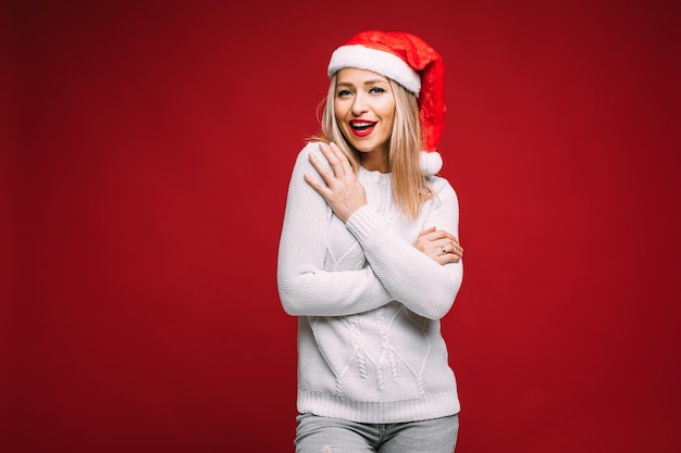 Foto di stock di attraente donna bionda con cappello da Babbo Natale rosso e maglione lavorato a maglia bianco che si abbraccia e sorride alla telecamera. Ritaglio su sfondo rosso.