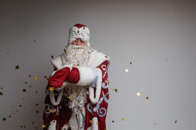 Foto di stock di jolly Father Frost in abiti festosi con una lunga barba bianca che allunga le braccia e sorride alla telecamera sotto i coriandoli dorati volanti. Isolato su sfondo grigio.