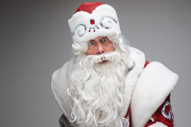 Foto di stock di stupito Babbo Natale in costume di fantasia con barba lunga bianca che guarda l'obbiettivo con occhi spalancati. Isolare su grigio.