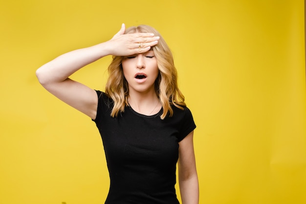 Foto di stock di una bionda donna caucasica in maglietta nera che tiene la mano sulla fronte con la bocca aperta e gli occhi chiusi. Dimostrazione di mal di testa. Donna che soffre di mal di testa. Sfondo giallo.