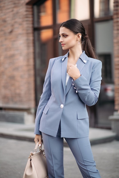 Foto di stock ritratto di attraente signora bruna elegante con coda in abito blu grigiastro con borsa beige in piedi in strada.