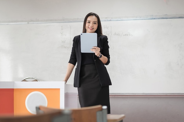 Foto di stock ritratto di un'insegnante donna asiatica allegra fiduciosa in un'uniforme nera con un tablet digitale e un laptop per insegnare la lingua moderna in classe