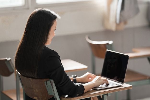 Foto di stock ritratto di un'insegnante donna asiatica allegra fiduciosa in un'uniforme nera con un tablet digitale e un laptop per insegnare la lingua moderna in classe