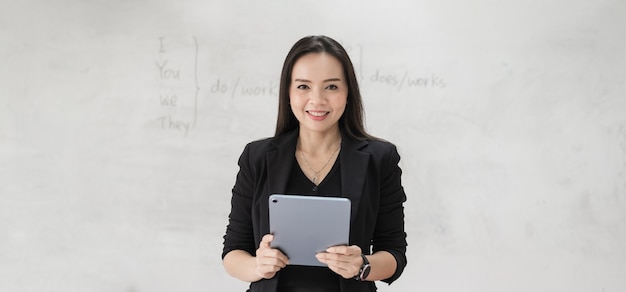 Foto di stock ritratto di un'insegnante donna asiatica allegra fiduciosa in un'uniforme nera con un tablet digitale e un laptop per insegnare la lingua moderna in classe