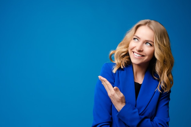 Foto di stock ritratto di una splendida bionda caucasica donna sorridente in giacca blu che mostra qualcosa con la sua mano. Copia spazio o spazio vuoto su sfondo blu brillante.