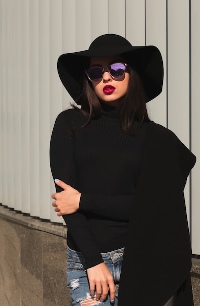 Foto di strada di una donna bruna affascinante in abbigliamento alla moda, indossa cappello e occhiali, posa alla luce del sole