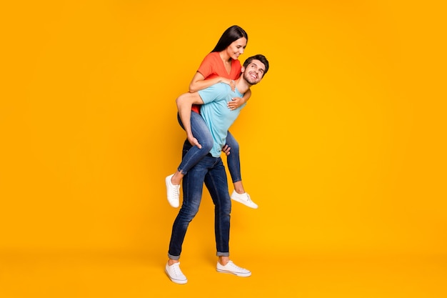 Foto di tutto il corpo di due persone divertenti ragazzo che trasportano lady piggyback incontrare avventure estive insieme indossare casual trendy blu arancione t-shirt jeans isolato muro di colore giallo