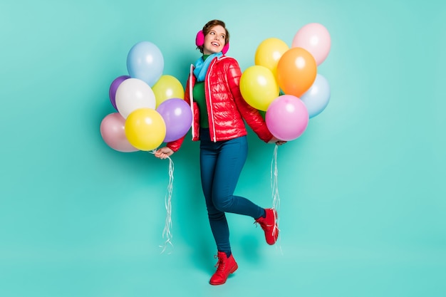 Foto di tutto il corpo di una fantastica festa di compleanno di una giovane donna porta palloncini colorati indossare casual cappotto rosso sciarpa rosa cuffie antirumore pantaloni scarpe vestito isolato muro color verde acqua